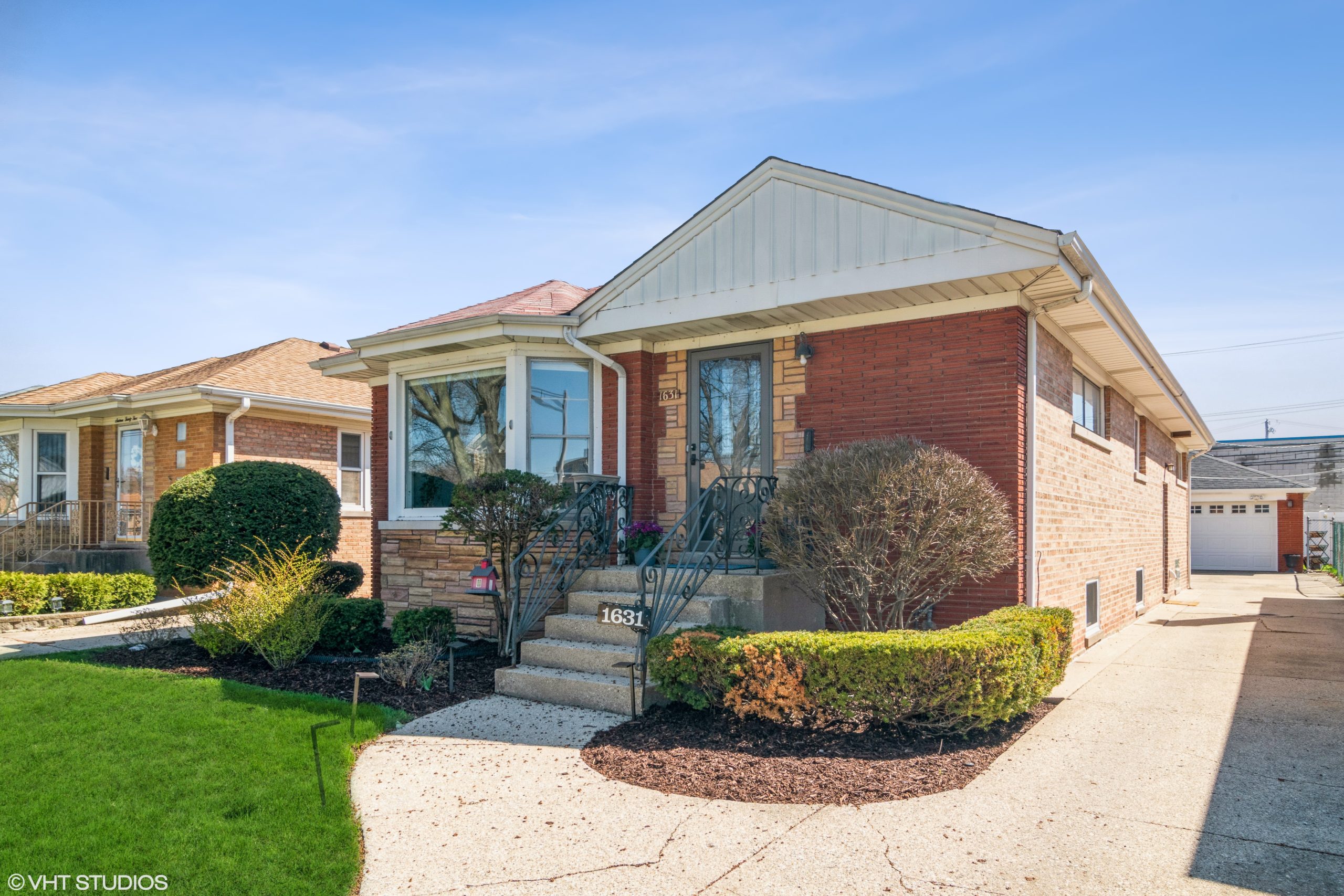 Mid-century brick ranch home with a front bay window, iron railings, and a small front stoop leading to a decorative glass door. The exterior features red and tan brickwork, neatly trimmed shrubs, and a wide concrete driveway leading to a detached garage in the rear. The home sits on a quiet, well-maintained suburban block.