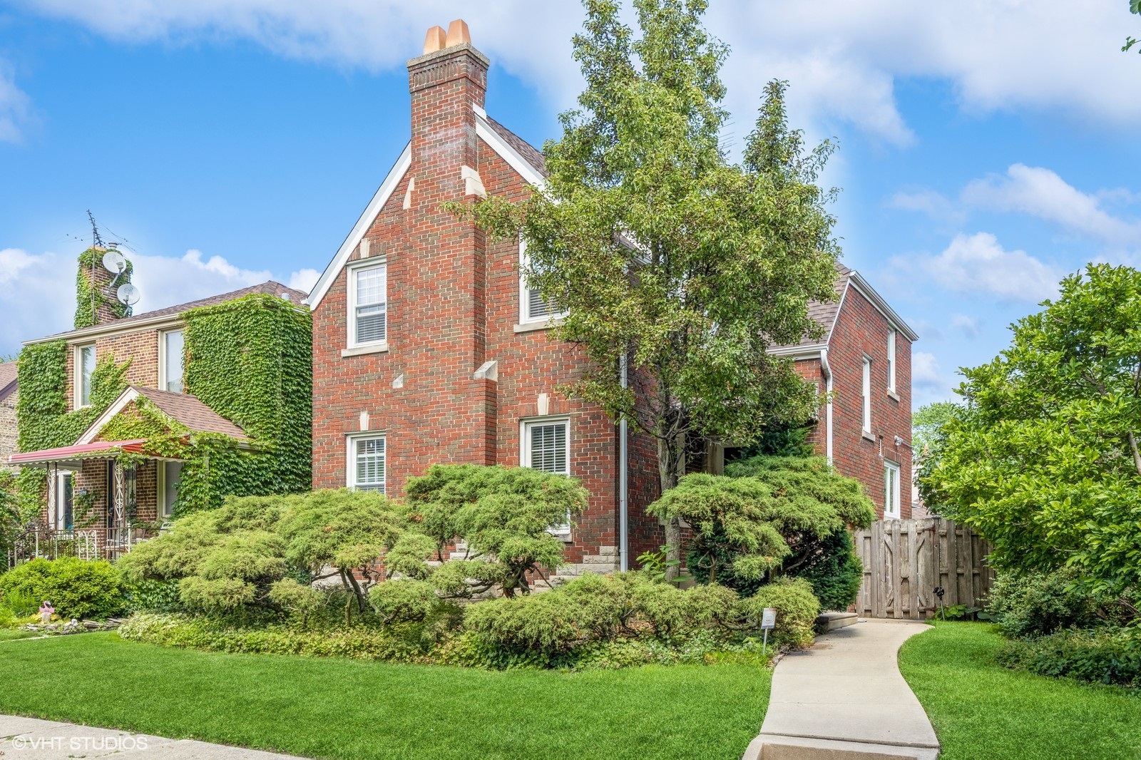 Charming brick Tudor-style home with a steep gabled roof, white stucco and wood trim detailing, and a red shingled roof. The home features a striped awning over the front bay window, manicured hedges, a tiered stone retaining wall, and a concrete walkway leading to the front steps. Neighboring homes in similar historic styles line the sunny, tree-lined street.