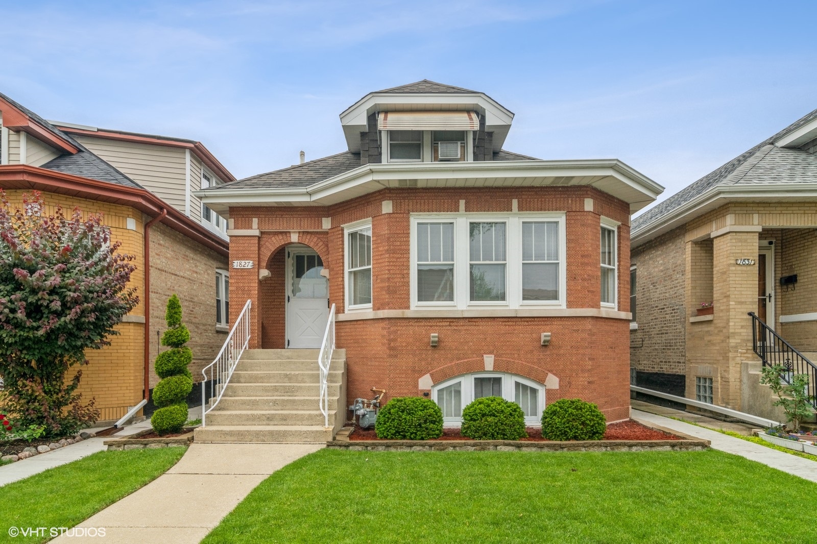 Classic Chicago brick bungalow with a raised front entrance, arched doorway, and prominent bay windows. The home features a small dormer with an awning, white trim accents, and manicured landscaping with neatly shaped bushes and a concrete walkway leading to the front steps. Flanked by similar historic homes on a residential block.