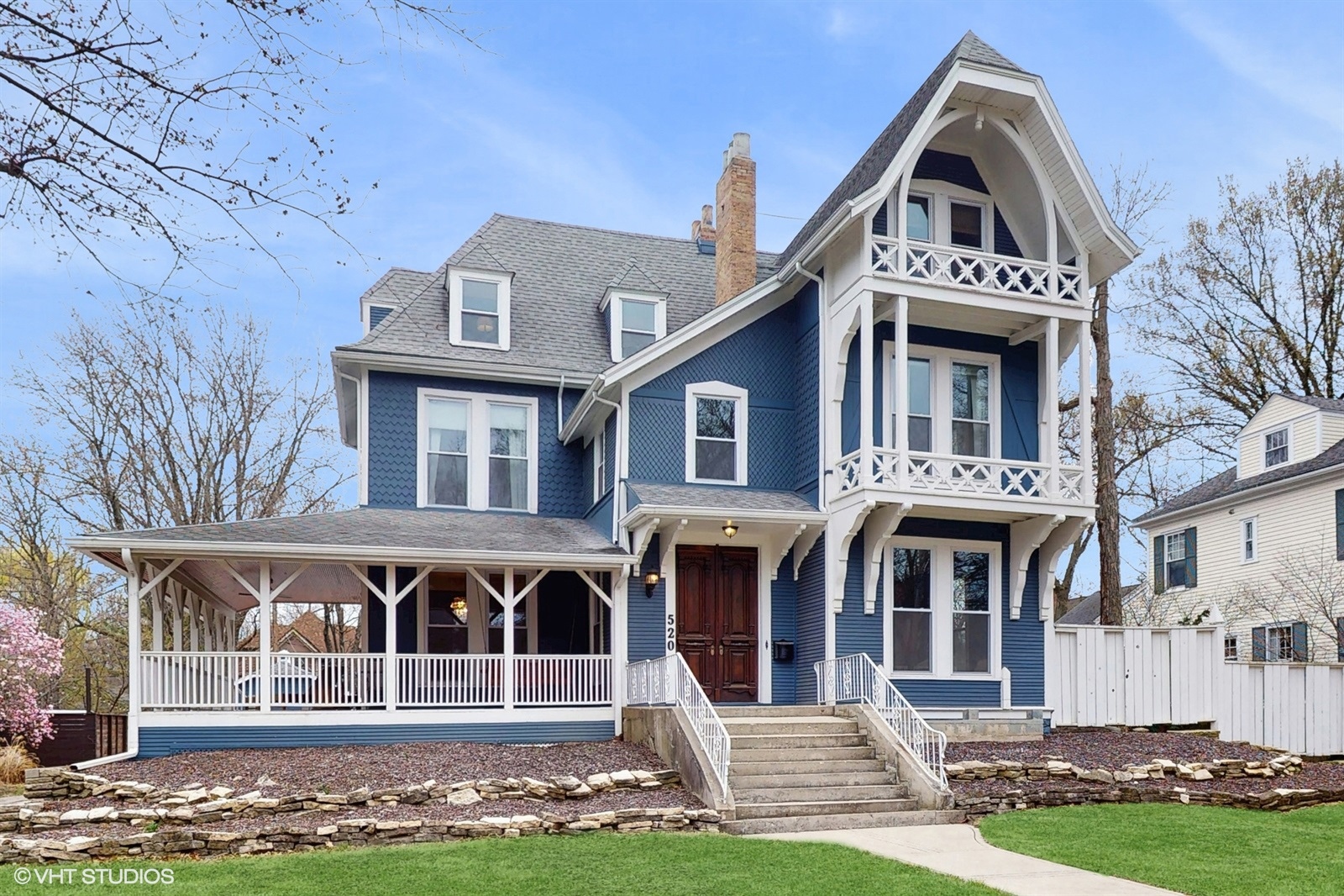 Victorian-style blue and white three-story home with a steep gabled roof, decorative trim, and a large wraparound porch. The home features ornate woodwork, a second-story balcony with lattice detailing, and a wide front staircase leading to double wooden doors. Spring trees and manicured lawn frame the property.