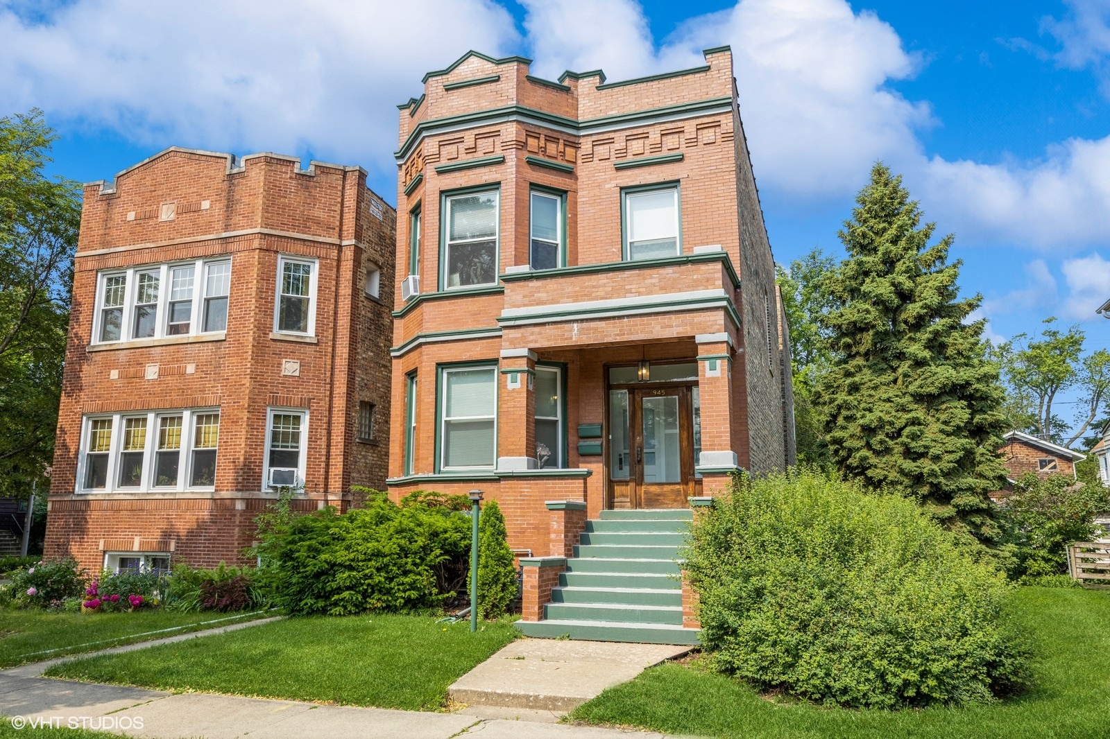 Classic two-flat red brick building with a flat roof, decorative stone detailing, and green-trimmed windows. The entry features a covered front porch with stairs leading to a wood-framed glass door. Well-maintained landscaping surrounds the home, which sits on a quiet residential street next to another similar historic structure.